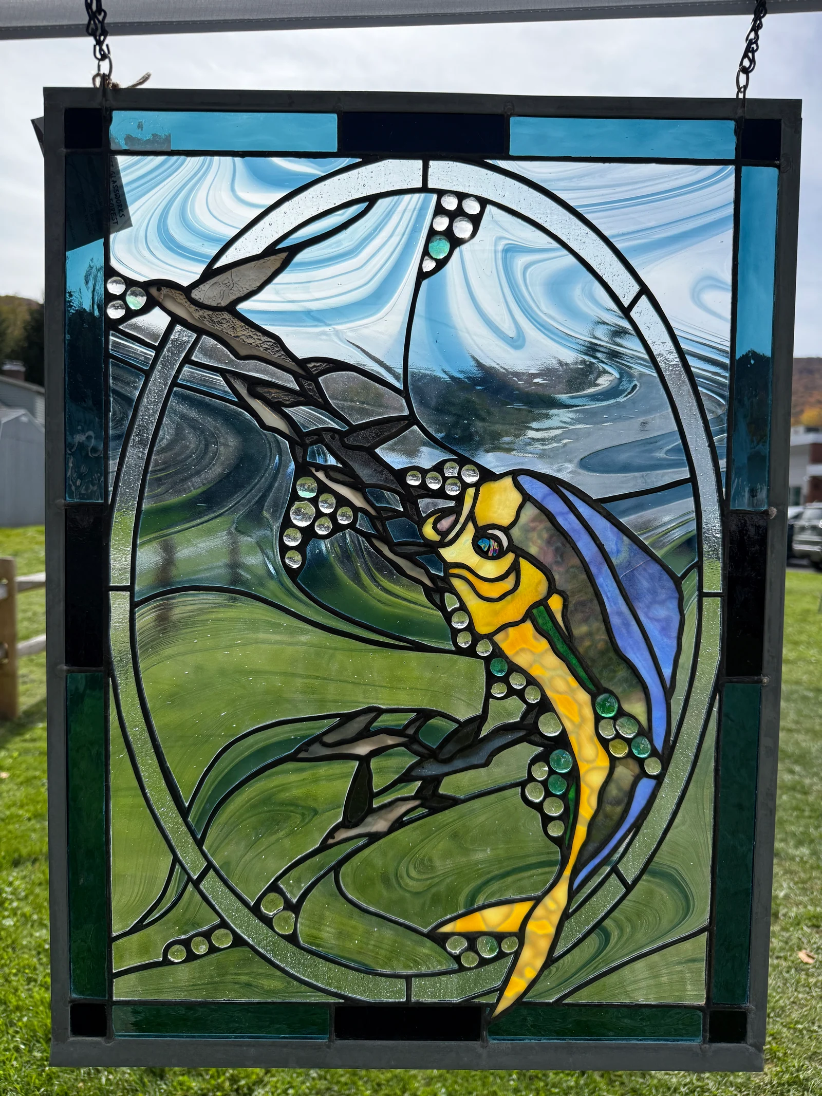 Golden fish stained-glass panel photographed outdoors with green grass behind the glass